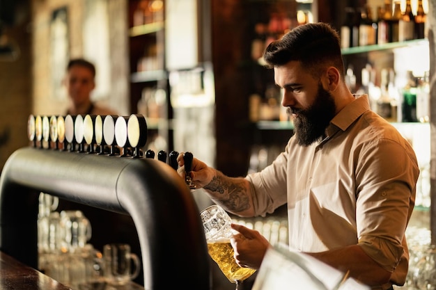 Jonge barman die tapbier in bierglas giet terwijl hij aan de toog werkt.