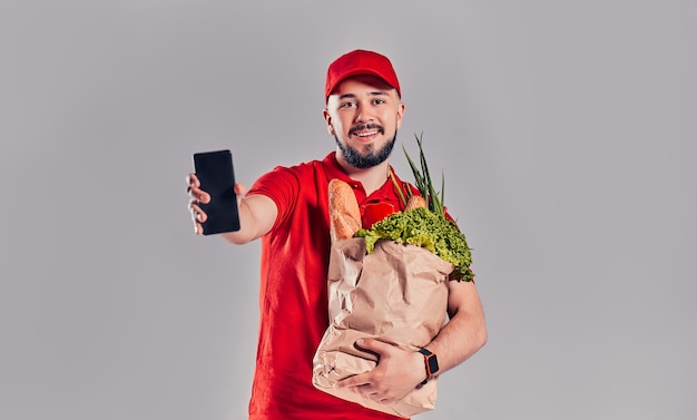 Jonge, bebaarde bezorger in rood uniform houdt pakket met brood en groenten vast en toont smartphonescherm geïsoleerd op grijze achtergrond.