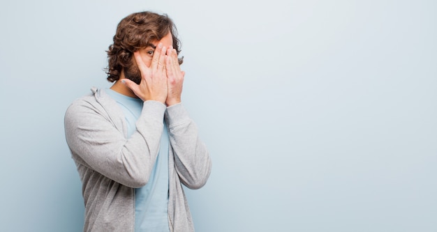 Jonge, bebaarde gekke man die gezicht bedekt met handen, gluren tussen vingers met verbaasde uitdrukking en kijken naar de zijkant tegen vlakke kleur