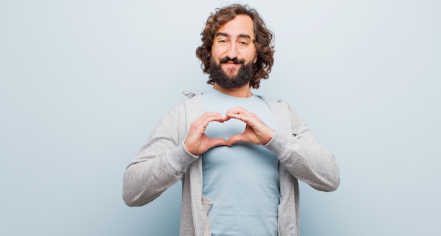 Jonge, bebaarde gekke man die lacht en zich gelukkig, schattig, romantisch en verliefd voelt, waardoor hij met beide handen een hartvorm krijgt