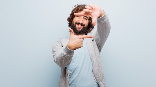 Jonge, bebaarde gekke man voelt zich gelukkig, vriendelijk en positief, glimlacht en maakt een portret of fotokader met handen tegen egale kleur