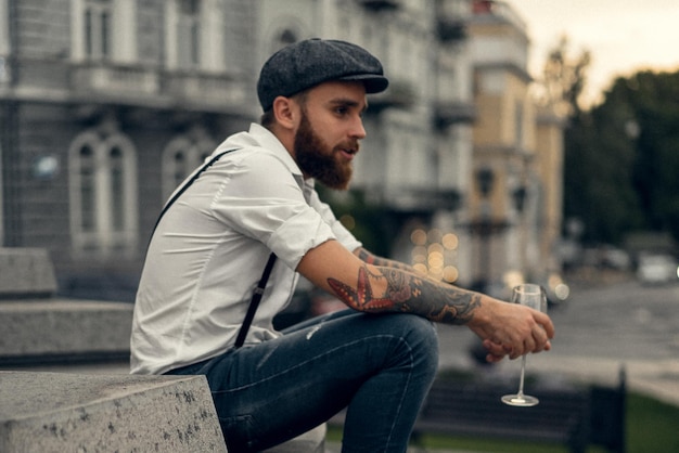 Jonge, bebaarde getatoeëerde man die wijn drinkt uit een glas op straat. Romantische man in een wit overhemd, pet en bretels in de stad. Peaky Blinders. ouderwets, retro.