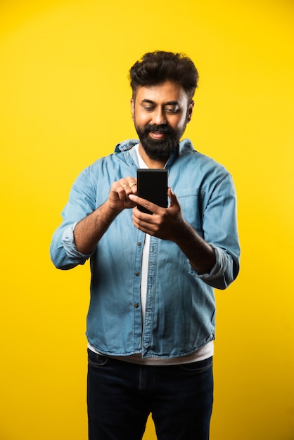 Jonge, bebaarde Indiase man die smartphone gebruikt, glimlacht tijdens het bellen of chatten met een vriend, staande op geel