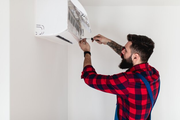 Jonge, bebaarde klusjesman die airconditioner repareert.