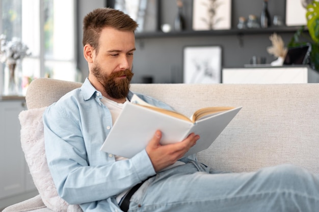 Foto jonge, bebaarde knappe man die thuis een boek leest op de bank