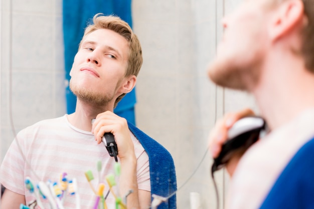 Foto jonge bebaarde knappe man met baard met machine in de badkamer in een spiegel kijken