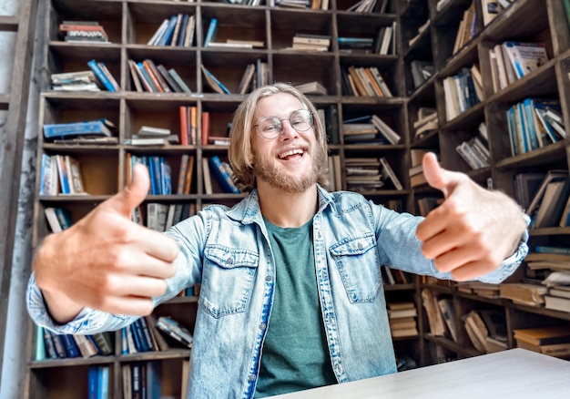 Jonge bebaarde lachende studentenblogger kijkt naar camera duimen omhoog