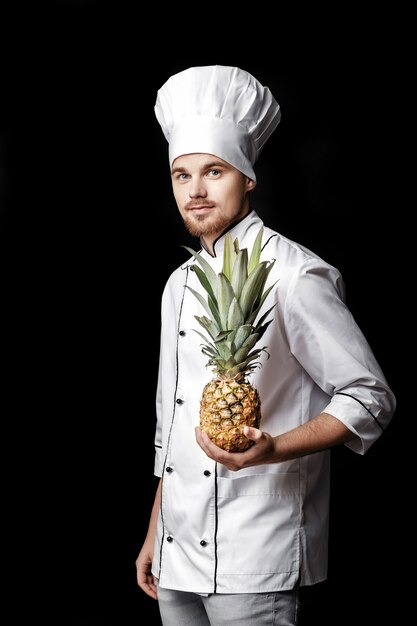 Jonge, bebaarde man chef-kok in wit uniform houdt verse ananas op zwarte achtergrond