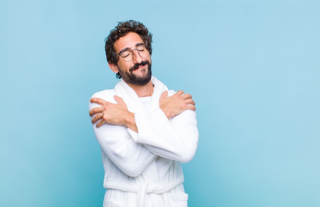 Jonge, bebaarde man die een badjas draagt die zich verliefd voelt en er schattig, schattig en gelukkig uitziet, romantisch lacht met de handen naast het gezicht