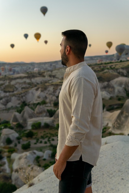 Jonge, bebaarde man die geniet van het uitzicht op heteluchtballonnen die bij zonsopgang vliegen in Cappadocië, Turkije