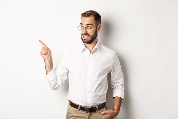 Jonge, bebaarde man die iets verontrustends ziet, ineenkrimpt terwijl hij met zijn vinger naar links wijst bij promo-aanbieding, onhandig staande tegen een witte achtergrond.