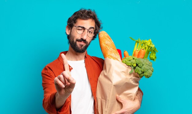 Foto jonge, bebaarde man die trots en zelfverzekerd glimlacht en nummer één triomfantelijk laat poseren, zich een leider voelt en een zak met groenten vasthoudt