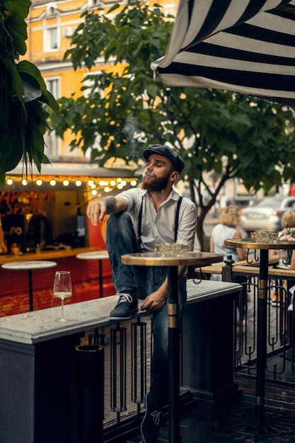 Jonge, bebaarde man in een café op straat met een glas wijn. Romantische man in een wit overhemd cap en bretels in de stad. Peaky Blinders. ouderwetse retro.