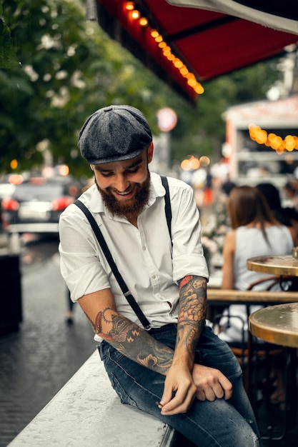 Jonge, bebaarde man in een café op straat rookt een sigaret. Romantische man in een wit overhemd cap en bretels in de stad. Peaky Blinders. oude modieuze retro.