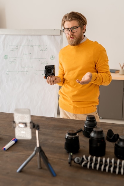 Jonge, bebaarde man met fotocamera legt uit hoe hij stockfoto's kan maken aan zijn online publiek voor de camera van de smartphone