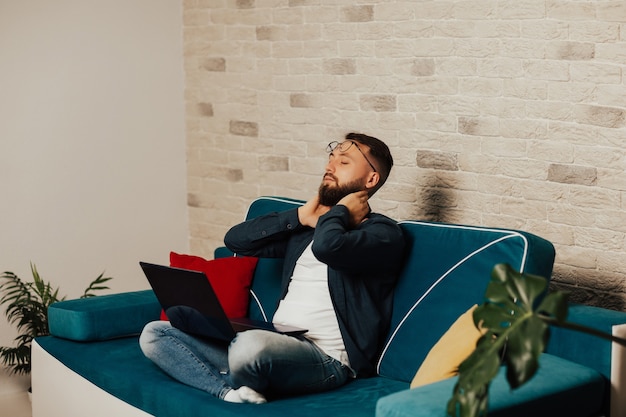 Jonge, bebaarde man ontspannen op een comfortabele bank met laptop. Rustige man lounge ogen gesloten ontspannen na hard werken op laptop.