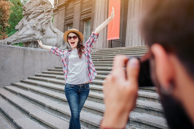 Jonge, bebaarde man praten foto van gelukkige vrouw. Ze poseert op camera. De vrouwelijke toerist houdt handen omhoog en draagt glazen. Ze staan op trappen.