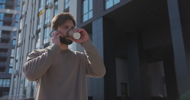 Jonge, bebaarde man praten op de smartphone en koffie drinken op straat Mensen en communicatieconcept