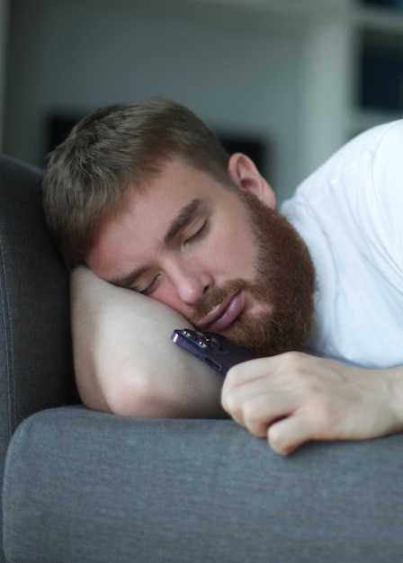 Jonge, bebaarde man slaapt thuis op de bank met zijn mobiele telefoon in de woonkamer smartphone