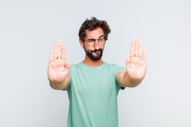 Jonge, bebaarde man ziet er serieus, ongelukkig, boos en ontevreden uit en verbiedt toegang of zegt stop met beide open handpalmen