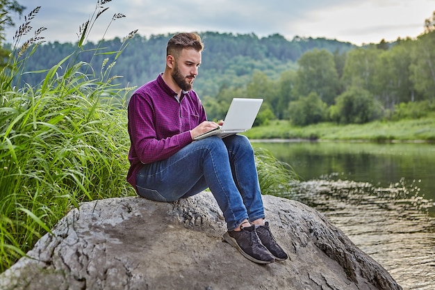 Jonge, bebaarde man zit op steen in de buurt van rivier met laptop op schoot, hij kijkt naar het scherm en typt tekst op het toetsenbord.