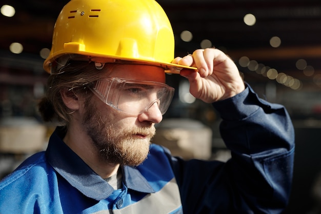 Jonge, bebaarde mannelijke ingenieur of fabrieksarbeider in beschermende helm en bril die voor de camera staat tijdens buitenwerk