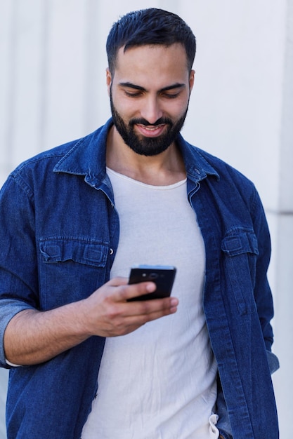 Jonge, bebaarde multiculturele man die voor een modern bedrijfsgebouw staat, glimlachend en typend op mobiele telefoon