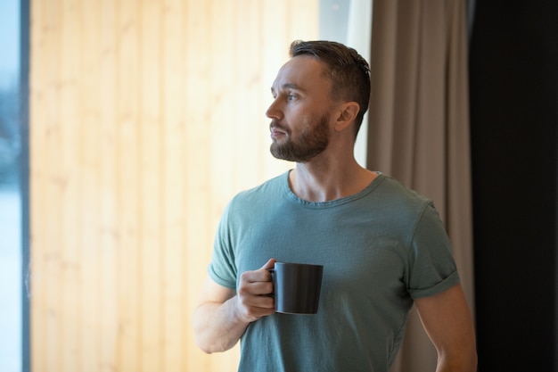 Jonge, bebaarde ontspannen man in grijs t-shirt met mok met hete thee of koffie en kijkend door een groot raam terwijl hij thuis blijft