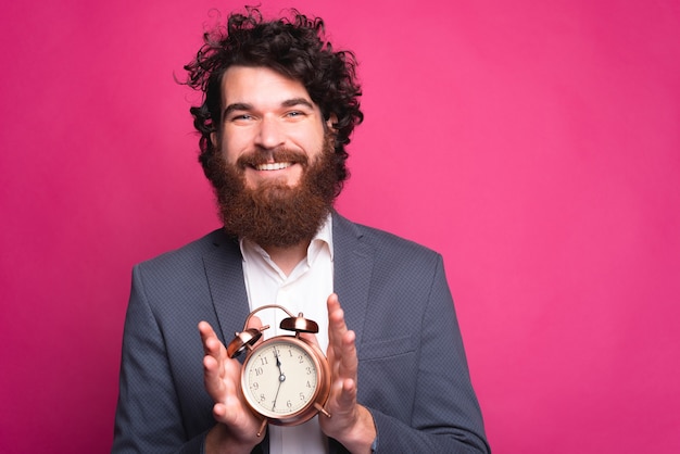 Jonge, bebaarde positieve man die een ronde klok houdt die en de camera glimlacht bekijkt