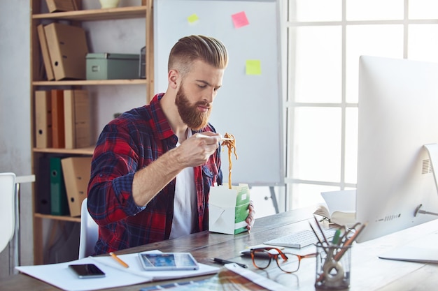 Jonge, bebaarde werknemer die voedsel eet terwijl hij aan het bureau zit in zijn moderne kantoor. Lifestyle-concept