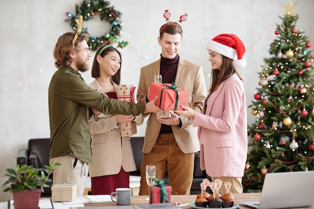 Jonge, bebaarde zakenman die een ingepakt kerstcadeau geeft aan vrouwelijke collega in kerstmuts terwijl hij haar feliciteert met kerstfeest op kantoor