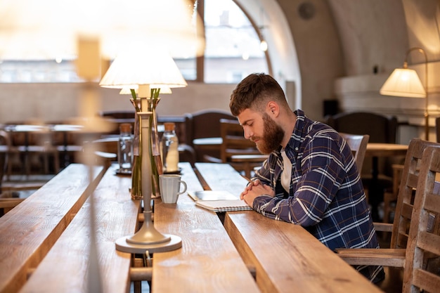 Jonge, bebaarde zakenman zit in café thuis aan tafel en schrijft in notitieboekje in de buurt van leugens tabletcomputer...