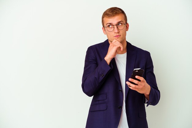 Jonge bedrijfsmens die een mobiele telefoon houdt die op witte muur wordt geïsoleerd die zijwaarts met twijfelachtige en sceptische uitdrukking kijkt.