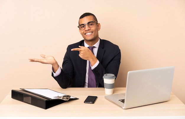 Jonge bedrijfsmens in zijn bureau met laptop en andere documenten die copyspace denkbeeldig op de palm houden om een advertentie op te nemen