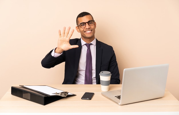 Jonge bedrijfsmens in zijn bureau met laptop en andere documenten die vijf met vingers tellen