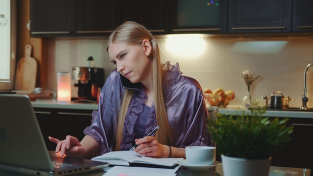 Jonge bedrijfsvrouw die thuis door bij de computer zit te werken en door smartphone te spreken