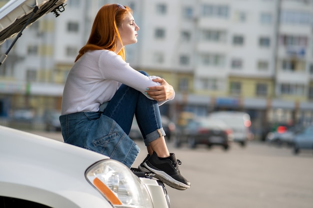 Jonge beklemtoonde vrouwenbestuurder dichtbij gebroken auto met geknalde kap die op hulp wacht.