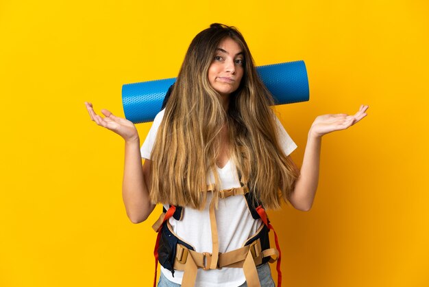 Jonge bergbeklimmervrouw met een grote rugzak die op gele muur wordt geïsoleerd die twijfels heeft terwijl het opheffen van handen