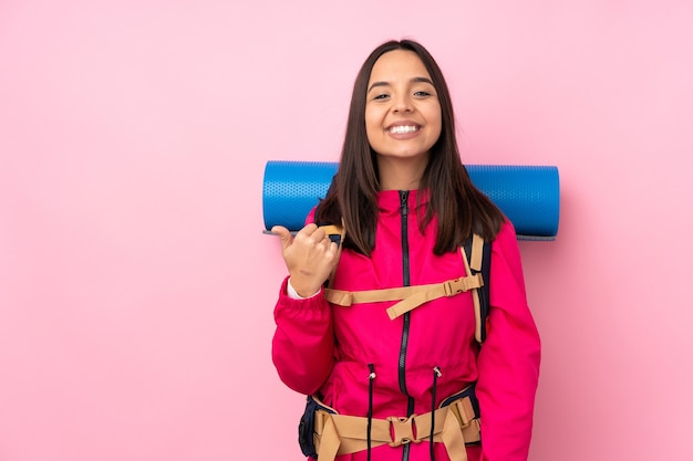 Jonge bergbeklimmervrouw met een grote rugzak op geïsoleerd roze die naar de kant wijst om een product te presenteren