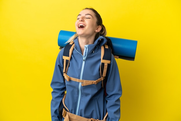 Jonge bergbeklimmervrouw met een grote rugzak over geïsoleerde gele achtergrond lachen