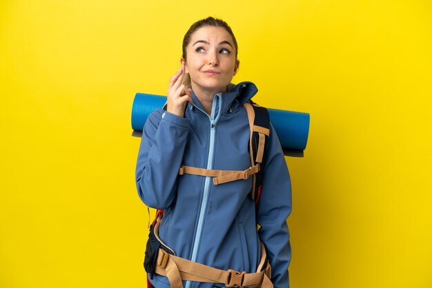 Jonge bergbeklimmervrouw met een grote rugzak over geïsoleerde gele achtergrond met vingers die kruisen en het beste wensen