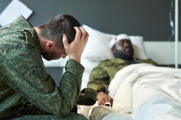 Jonge bezorgde soldaat in camouflage die hoofd aanraakt terwijl hij bij zijn vriend zit
