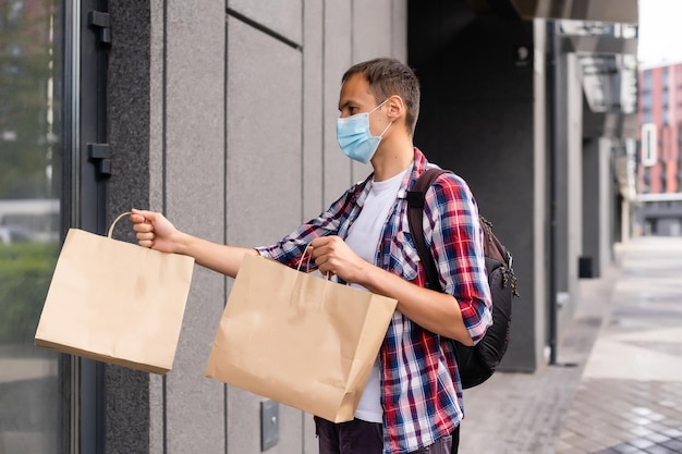 Jonge bezorger met medisch masker op gezicht met papieren zakken in handen. Hipster man koerier met boodschappentassen. Veilige online bezorgservice. Coronavirus COVID-19 bescherming