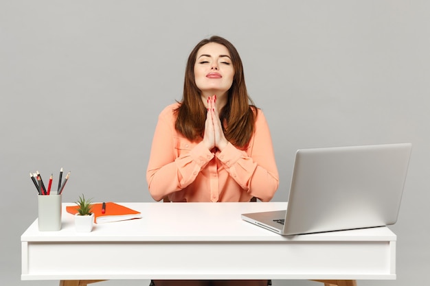 Jonge biddende vrouw in pastelkleren hand in hand gevouwen, ogen gesloten houden zitten, werken aan bureau met pc-laptop geïsoleerd op een grijze achtergrond. Prestatie zakelijke carrière concept. Bespotten kopie ruimte.