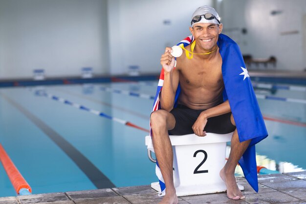 Foto jonge biraciale mannelijke zwemmer viert aan het zwembad gedrapeerd in een australische vlag met een medaille