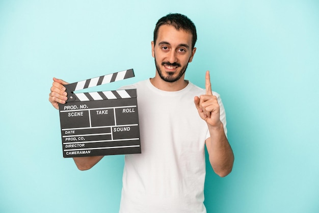 Jonge blanke acteur man met filmklapper geïsoleerd op blauwe achtergrond met nummer één met vinger.