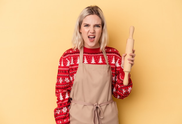 Jonge blanke bakkersvrouw die koekjes doet voor Kerstmis geïsoleerd op een gele achtergrond die erg boos en agressief schreeuwt.