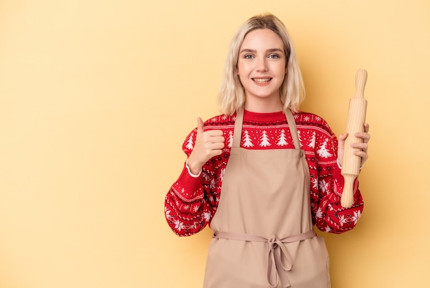 Jonge blanke bakkersvrouw die koekjes doet voor Kerstmis geïsoleerd op een gele achtergrond glimlachend en duim omhoog