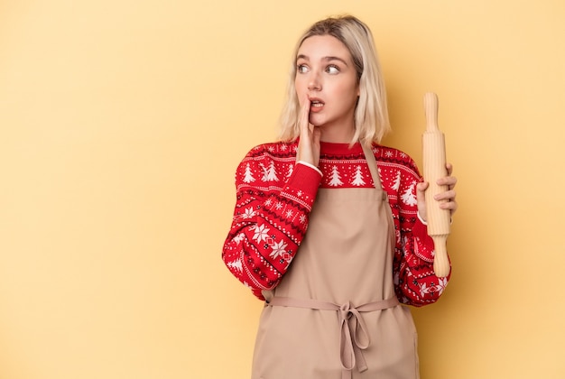 Jonge blanke bakkersvrouw die koekjes doet voor Kerstmis geïsoleerd op een gele achtergrond, zegt een geheim heet remnieuws en kijkt opzij