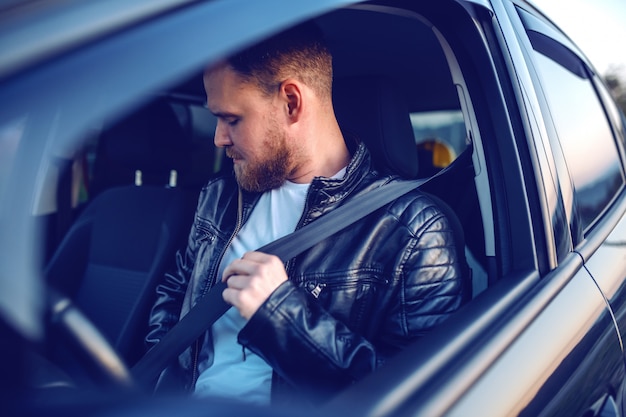 Jonge blanke bebaarde blonde man in lederen jas zit in zijn moderne auto en veiligheidsgordel. Veiligheid eerst.